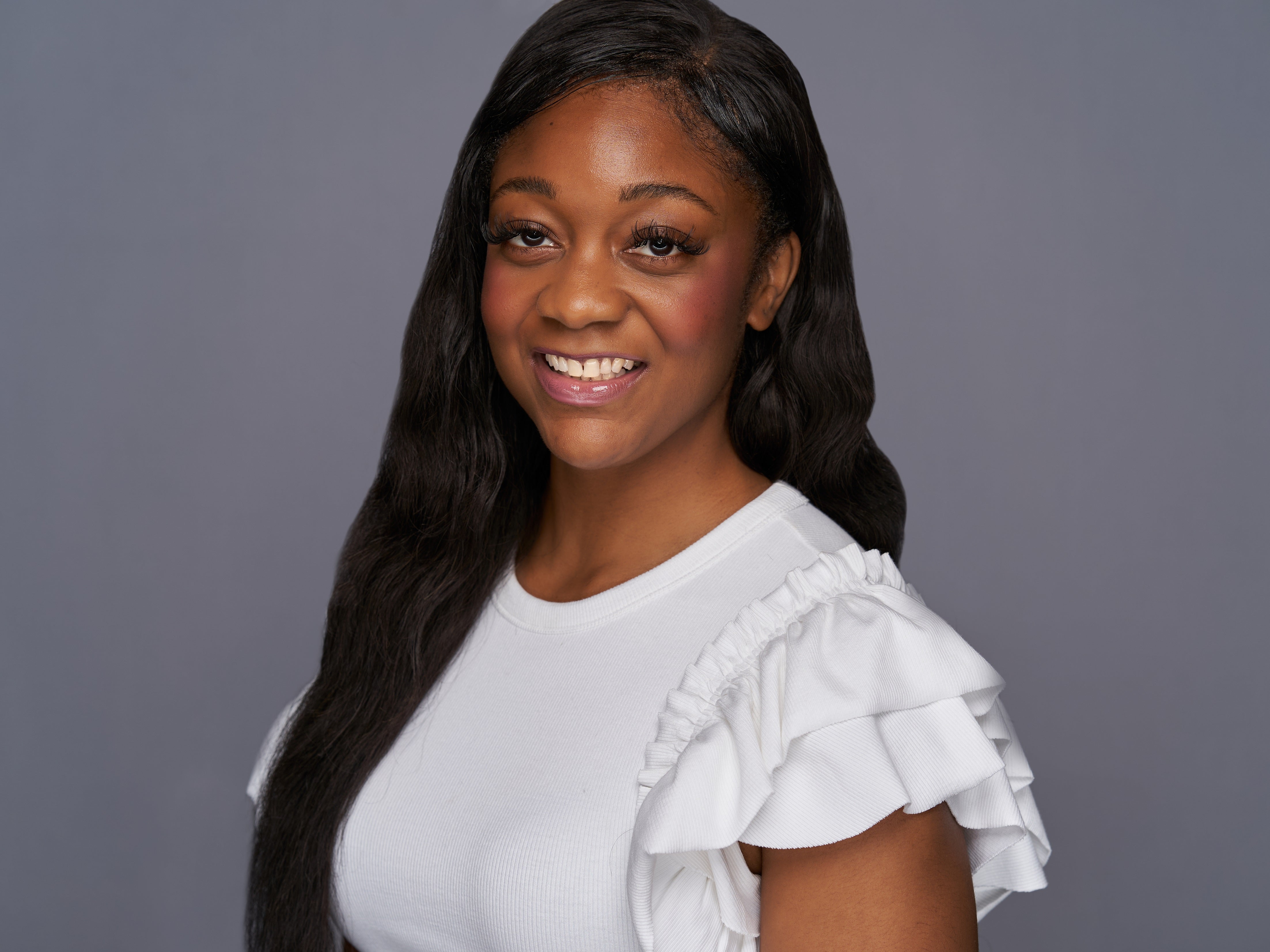 Shaina Welch poses for a headshot in a white blouse with her hair over one shoulder against a grey backdrop.