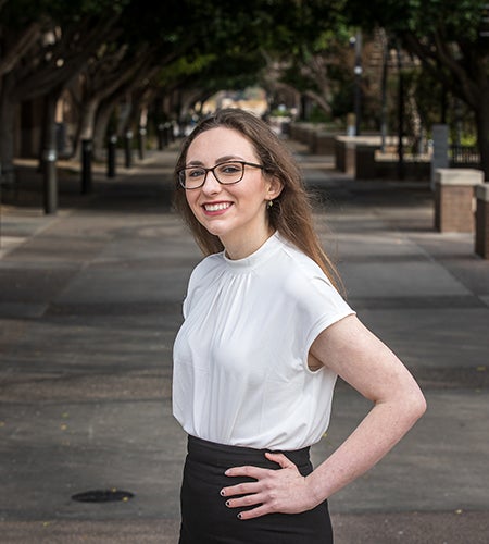 A female high school student in a white blouse poses for a photo in front of a long sidewalk lined with trees