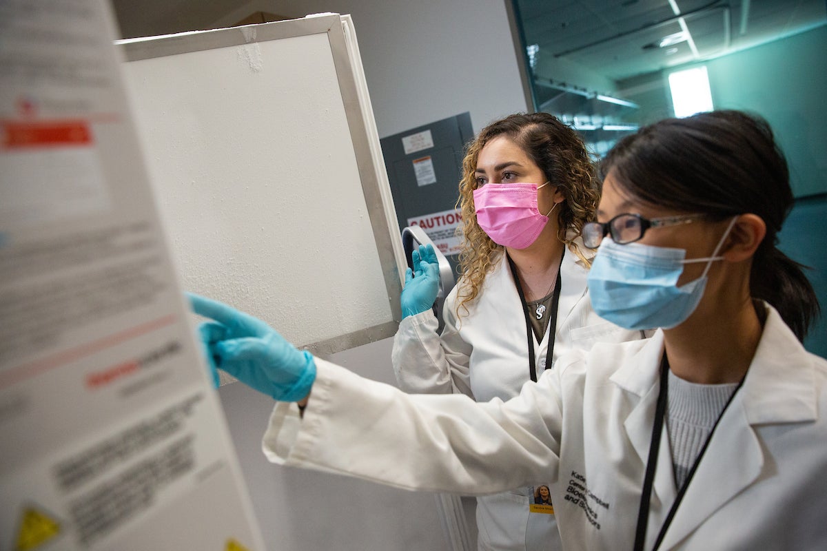 Students in lab coats, medical masks and gloves point at a chart