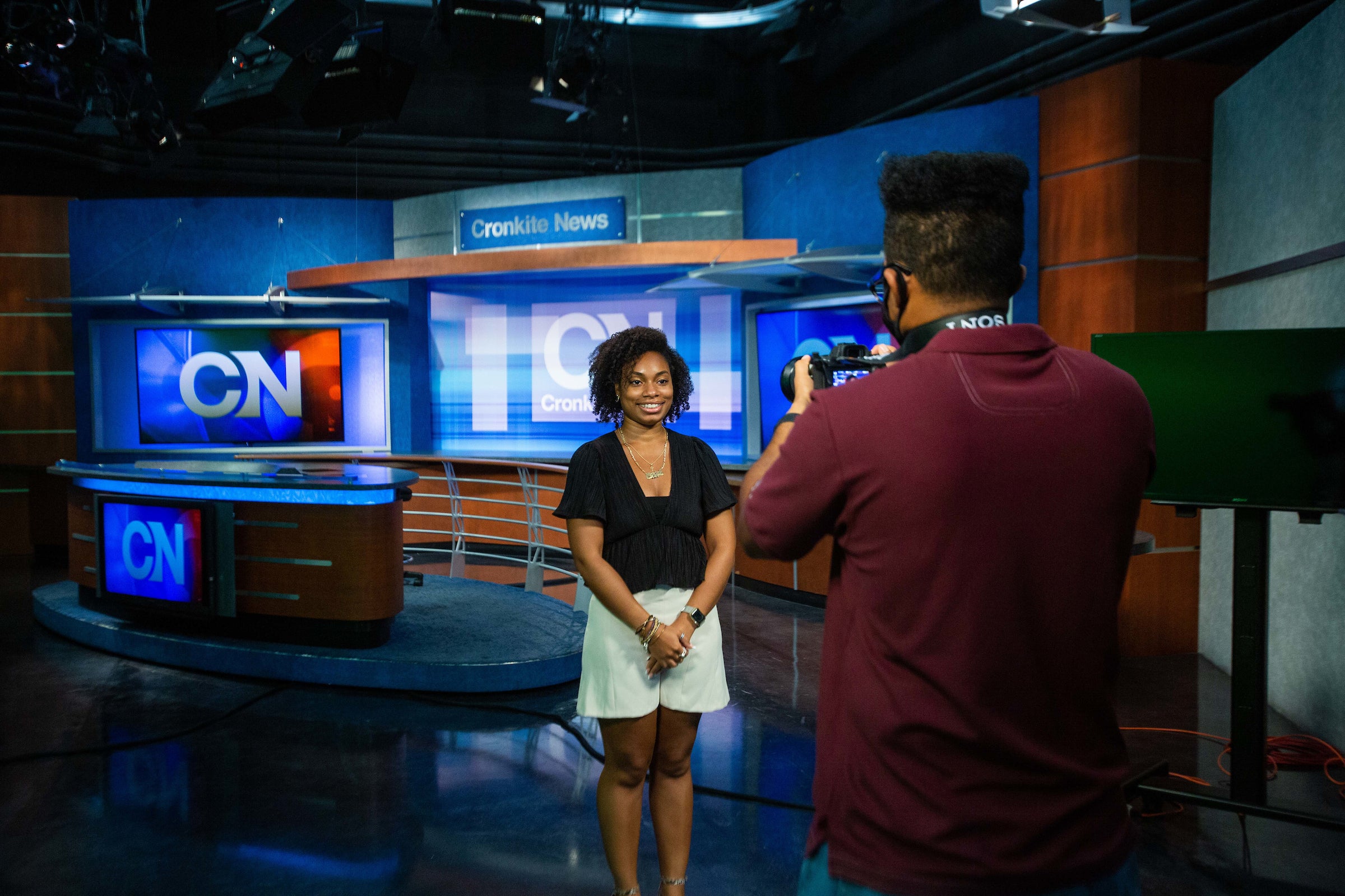 A college student smiles for a headshot with a news desk set behind her