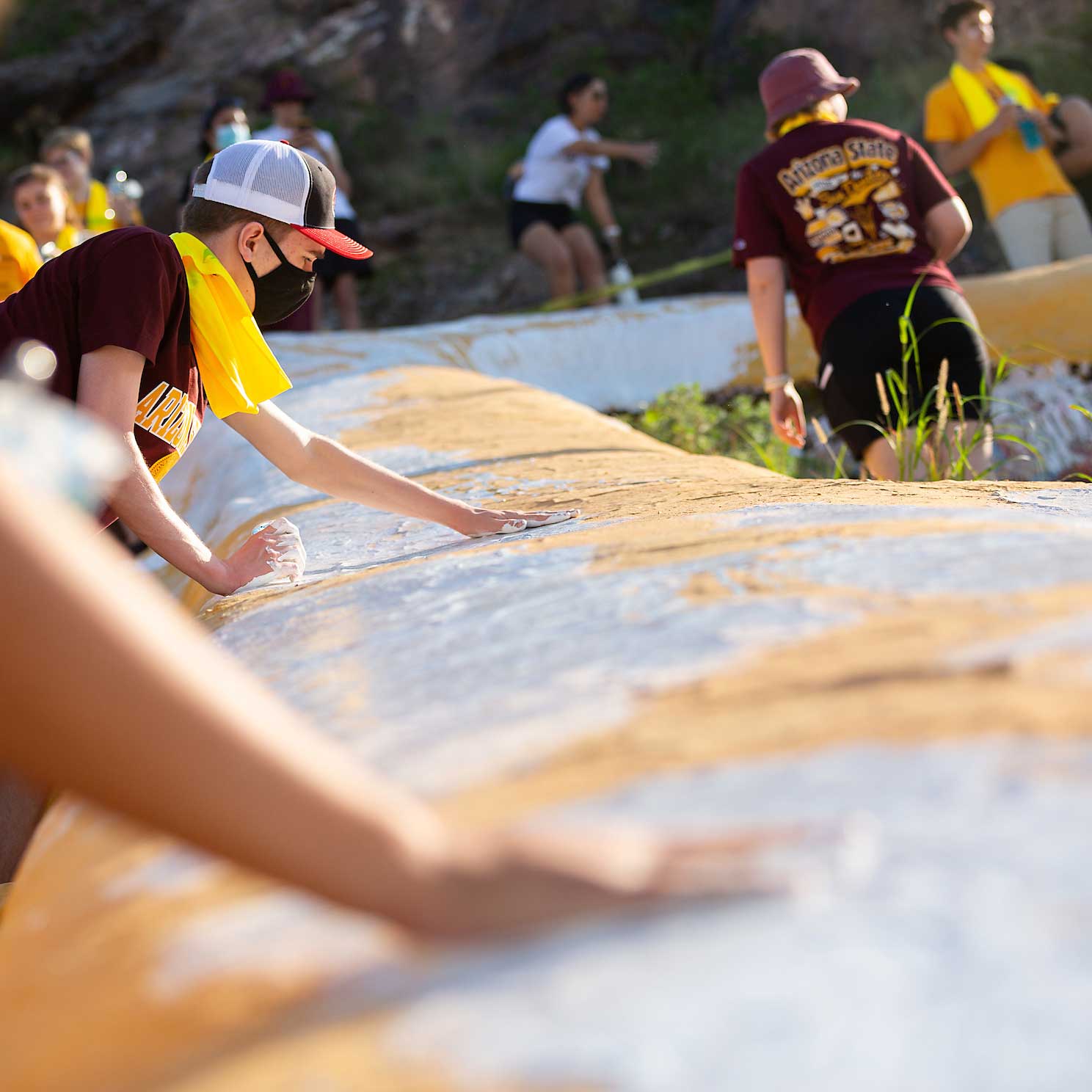 A student wearing a mask and ballcap spreads white paint on the A on A Mountain