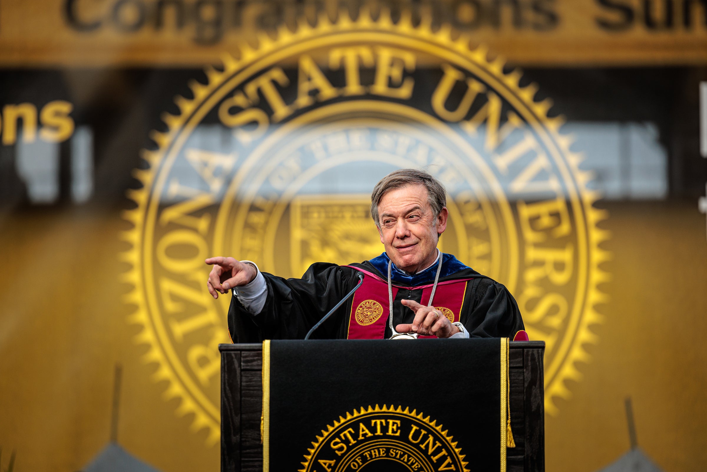 ASU President Crow stands at a lectern wearing graduation regalia and points to the audience while grinning