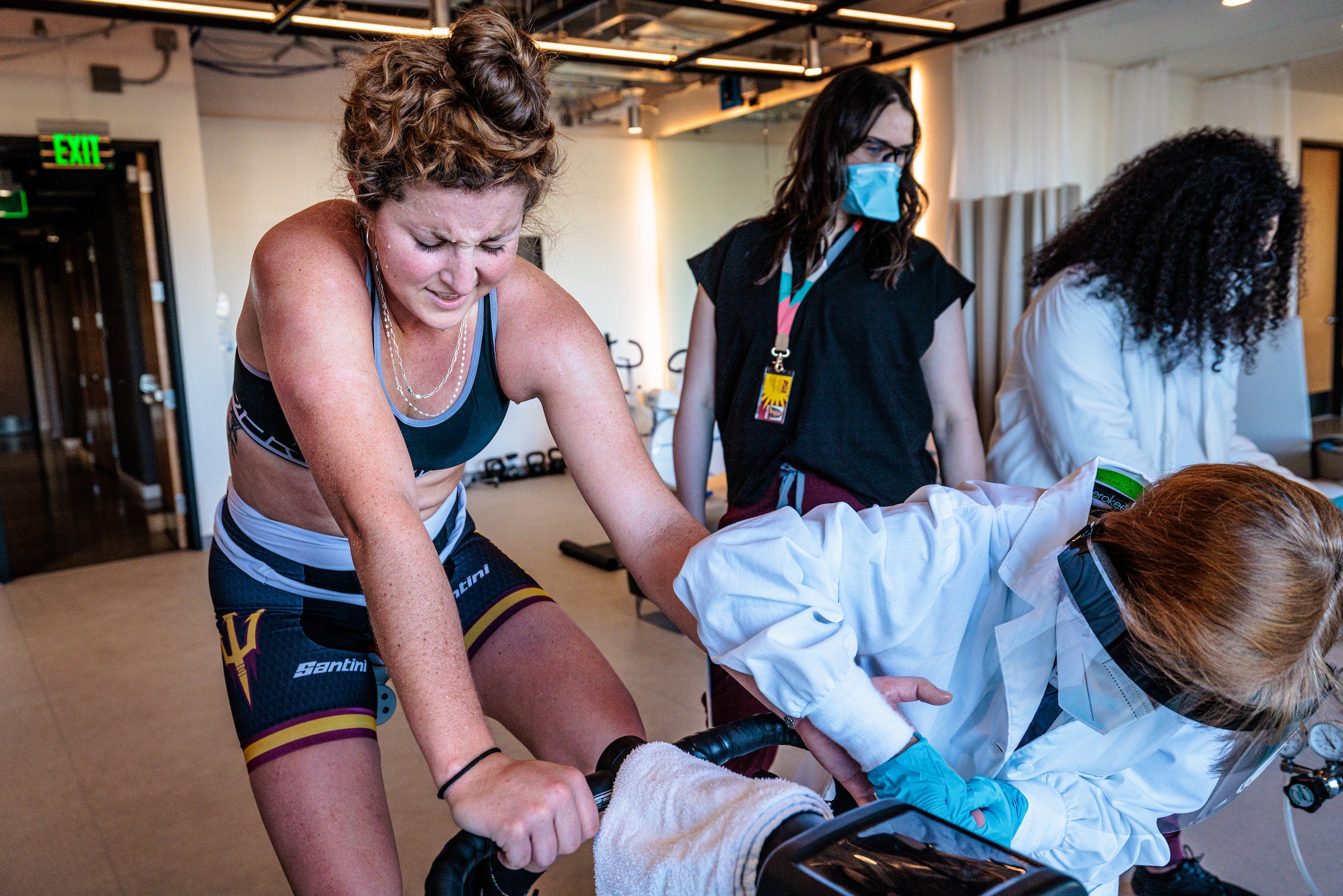 A woman with sweat pouring down her face as she cycles on a stationary bike grimaces as a phlebotomist takes a blood sample from her finger.