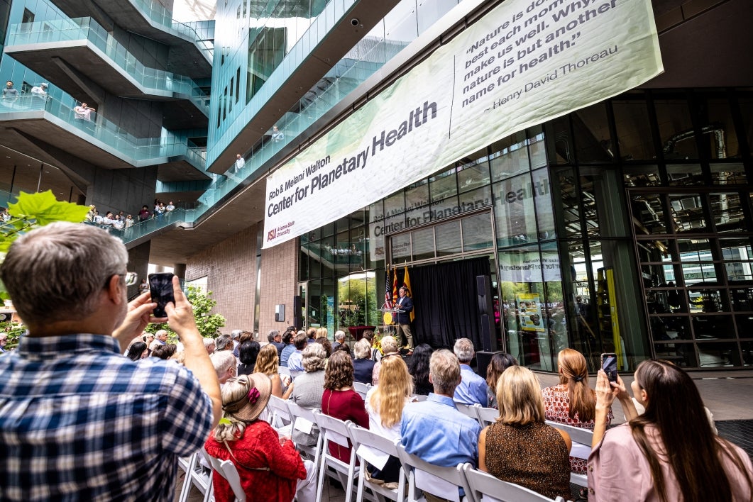 Audience members take phone photos of a sign hanging over a stage with the new name of the building, the Rob and Melani Walton Center for Planetary Health