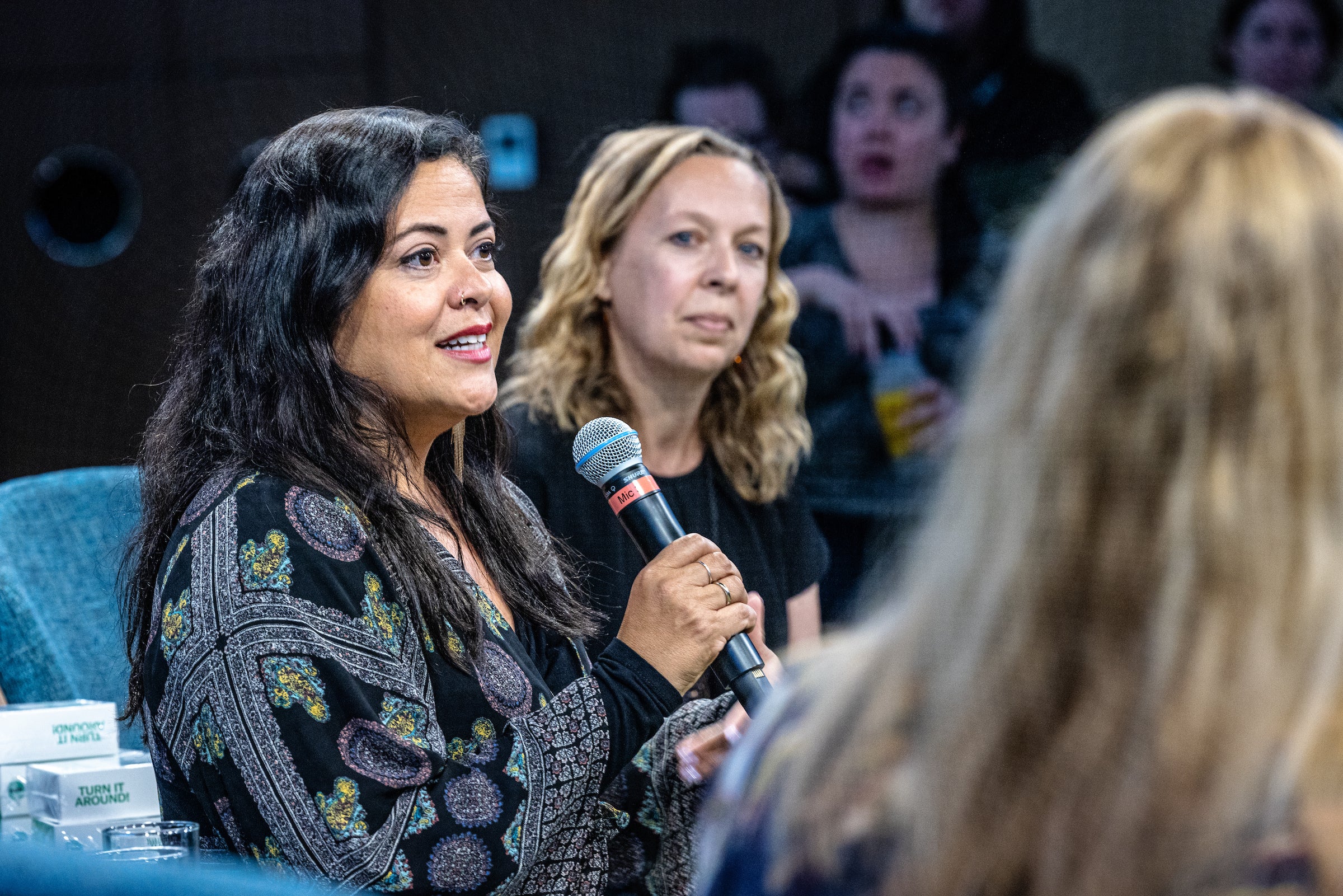 A woman speaks into a mic with other women seated around her