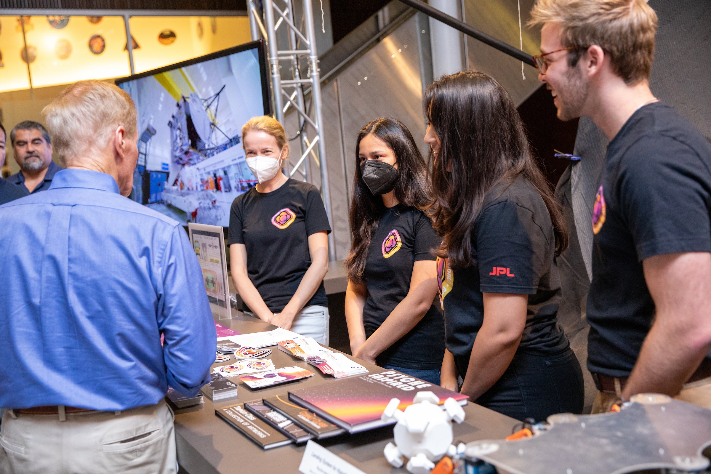 Students wearing Psyche team shirts stand behind a table with various mission items on it