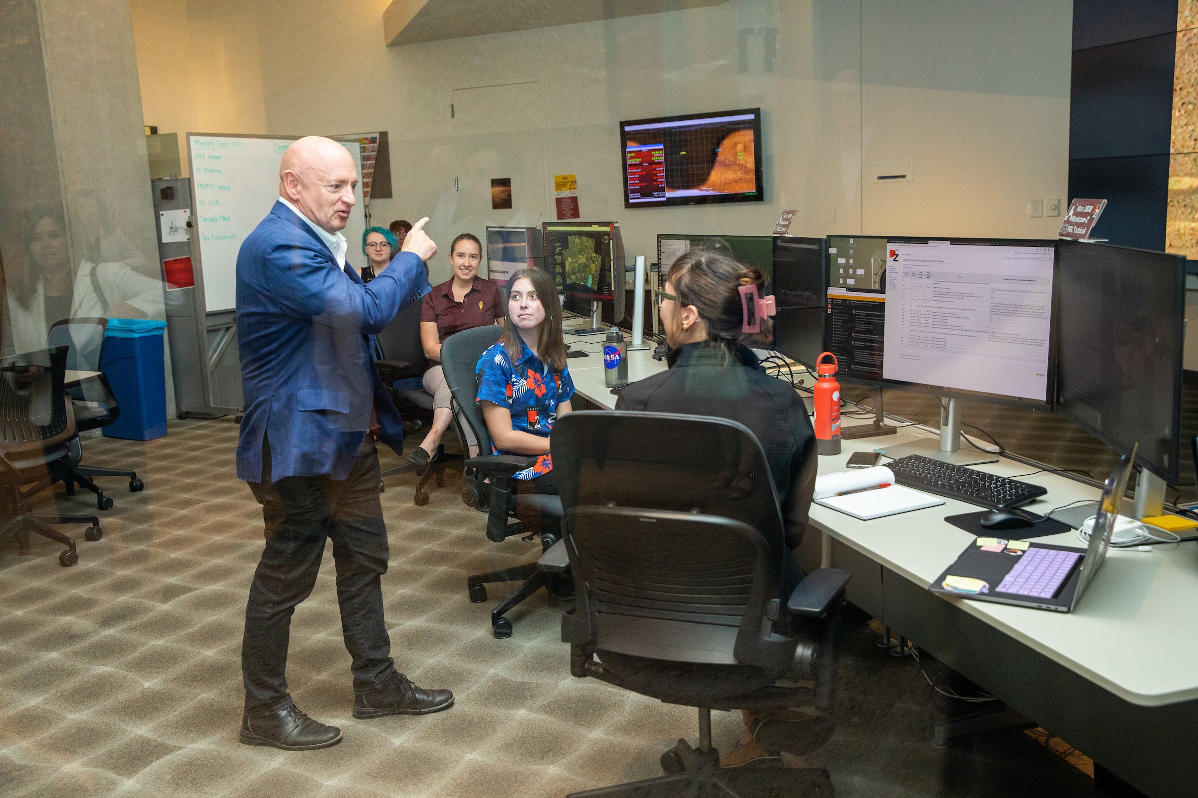 A man stands and talks to students seated in front of computer monitors