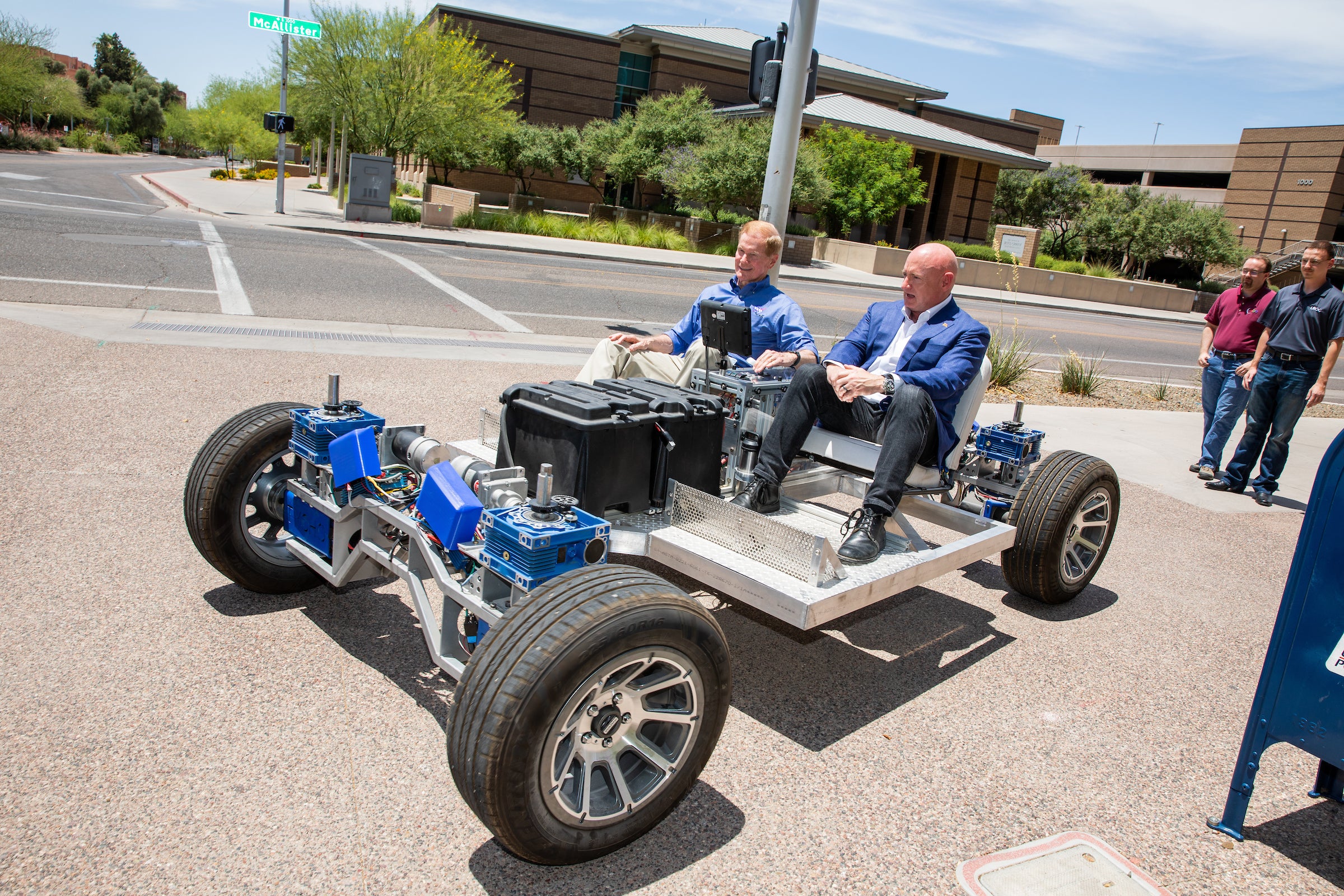 Two men sit in a lunar rover prototype on a sidewalk