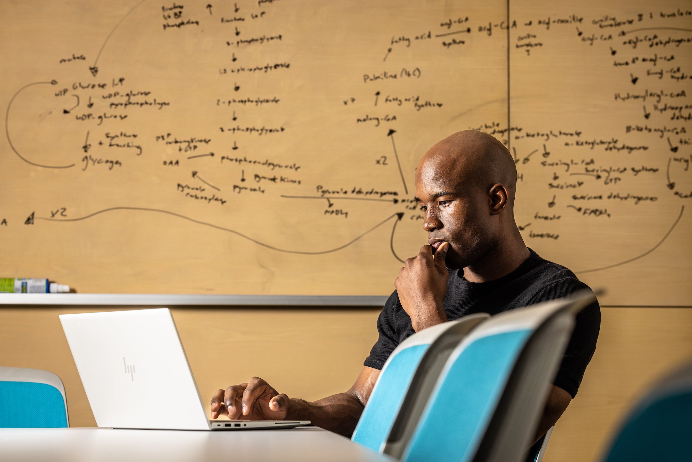 Man sitting at computer in classroom with writing on the whiteboard behind him 