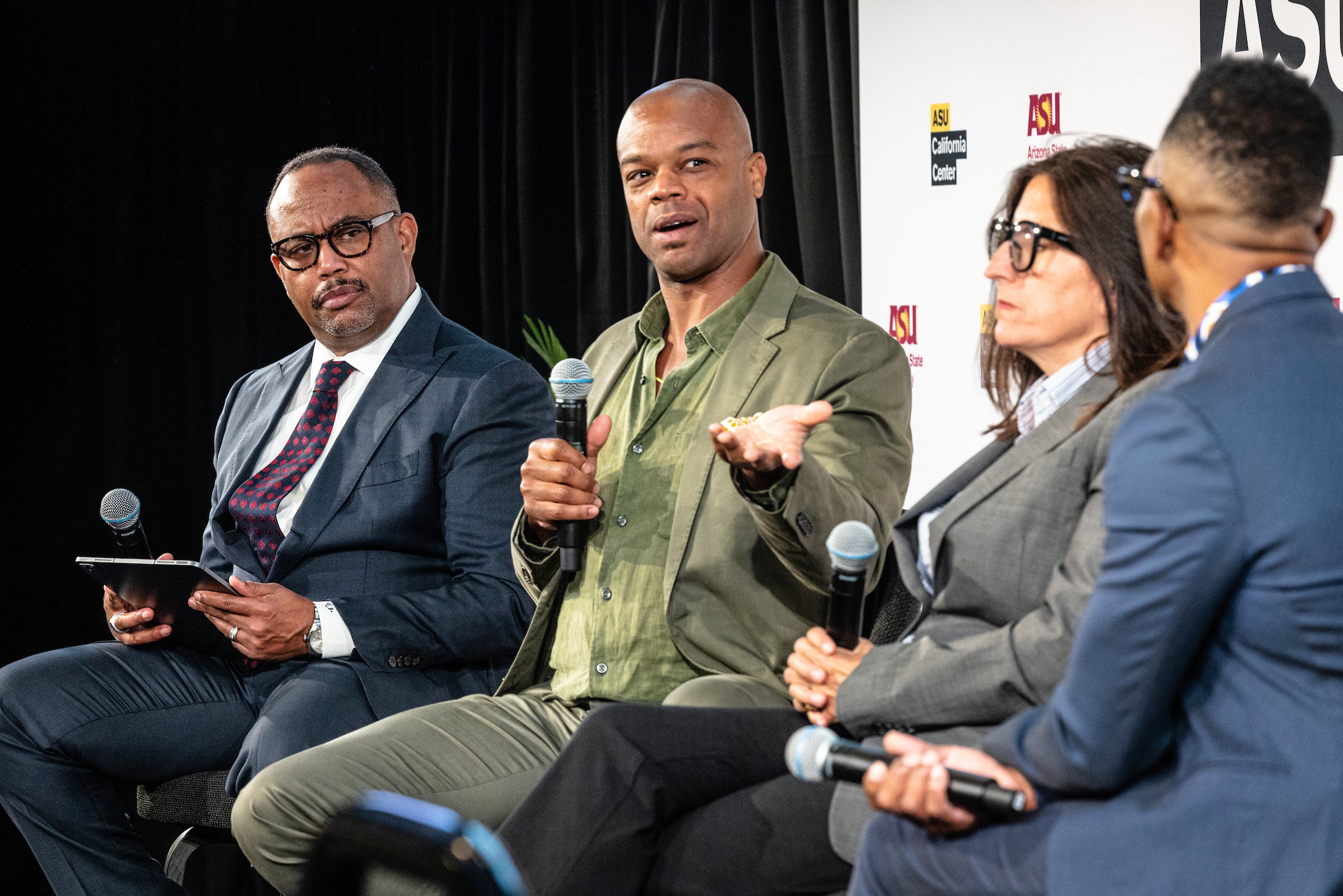 One man speaks as part of a panel while the other three panelists listen