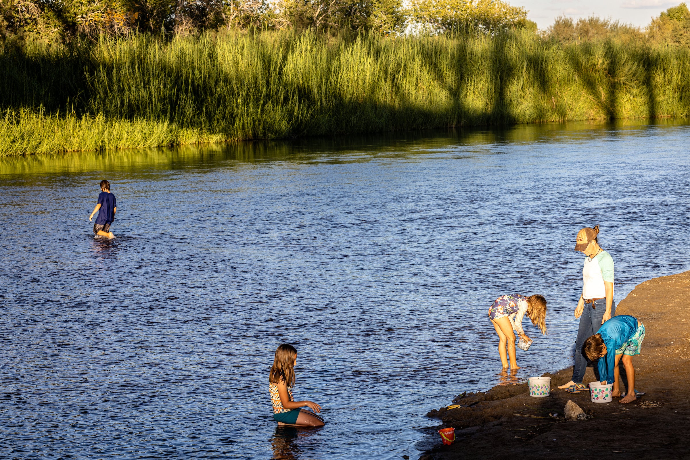 A mom watches her children as they play on the edge of the Colorado River and a teen wades through it