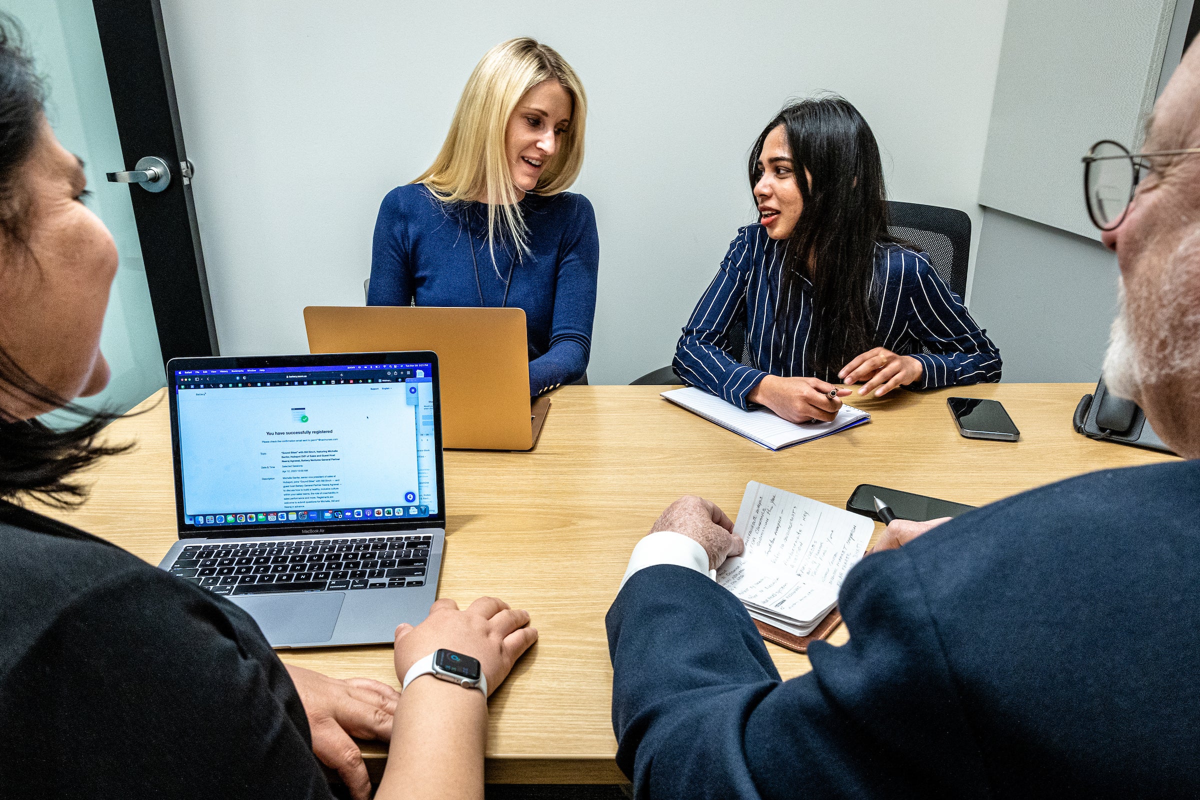 Group of four people sit around table with laptops and notebooks talking