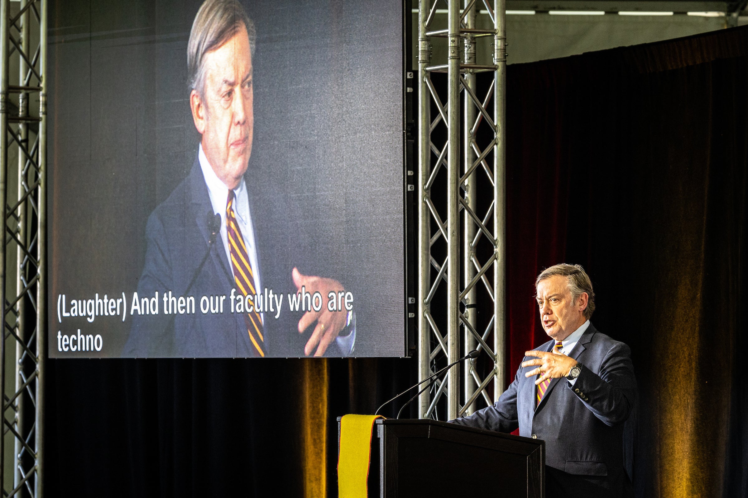 A man speaks at a lectern with his image projected on a large screen behind him