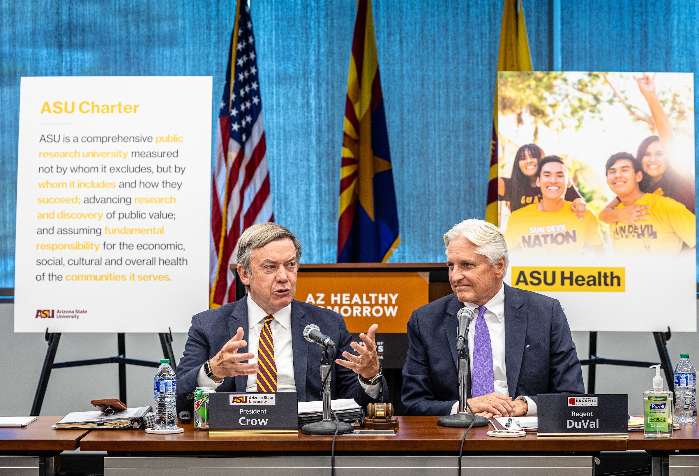 Two men speak at a conference table in front of signs and flags