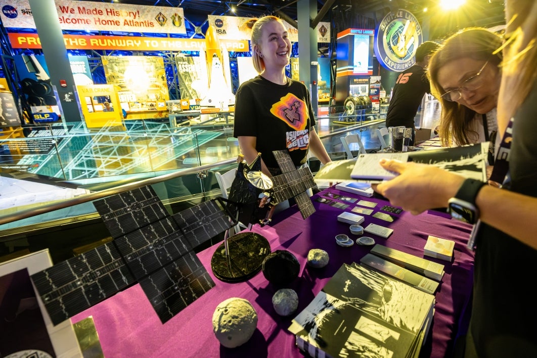 A college student speaks to visitors at a table covered in Psyche stickers and models