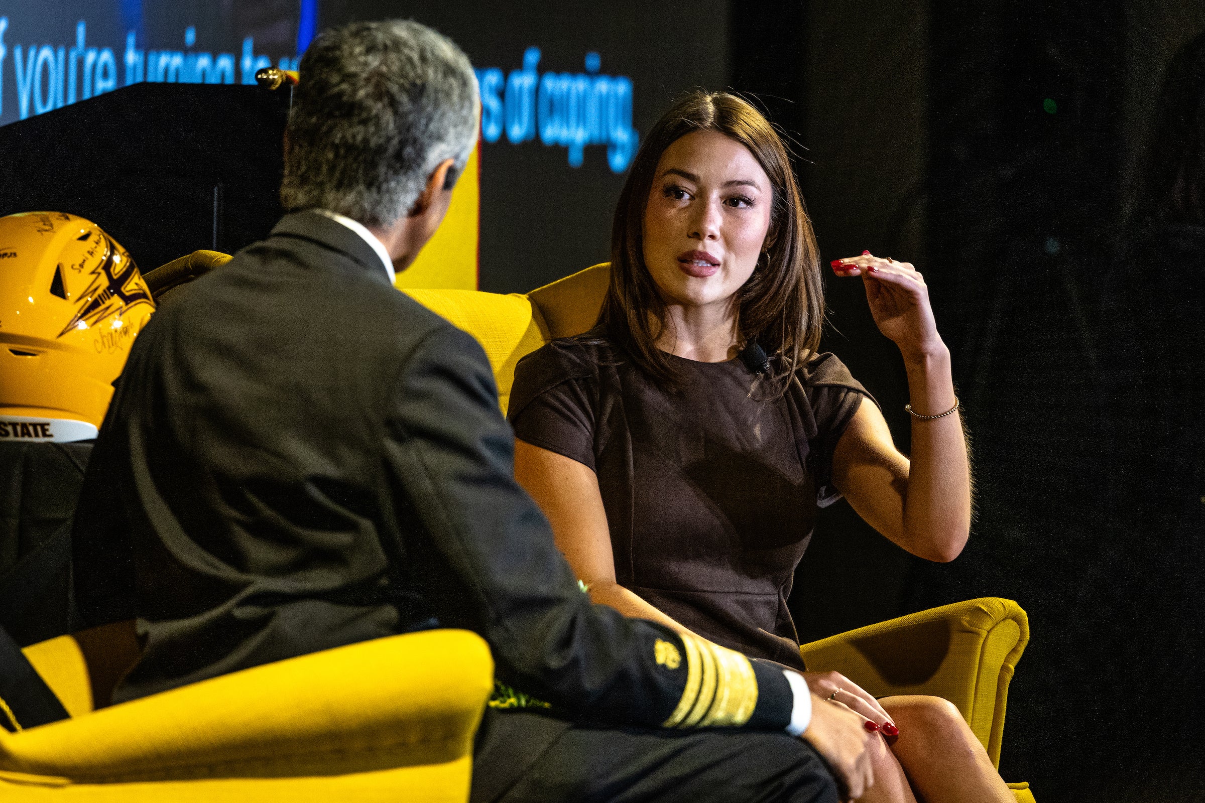 Young woman with long brown hair seated and talking to US surgeon general during event