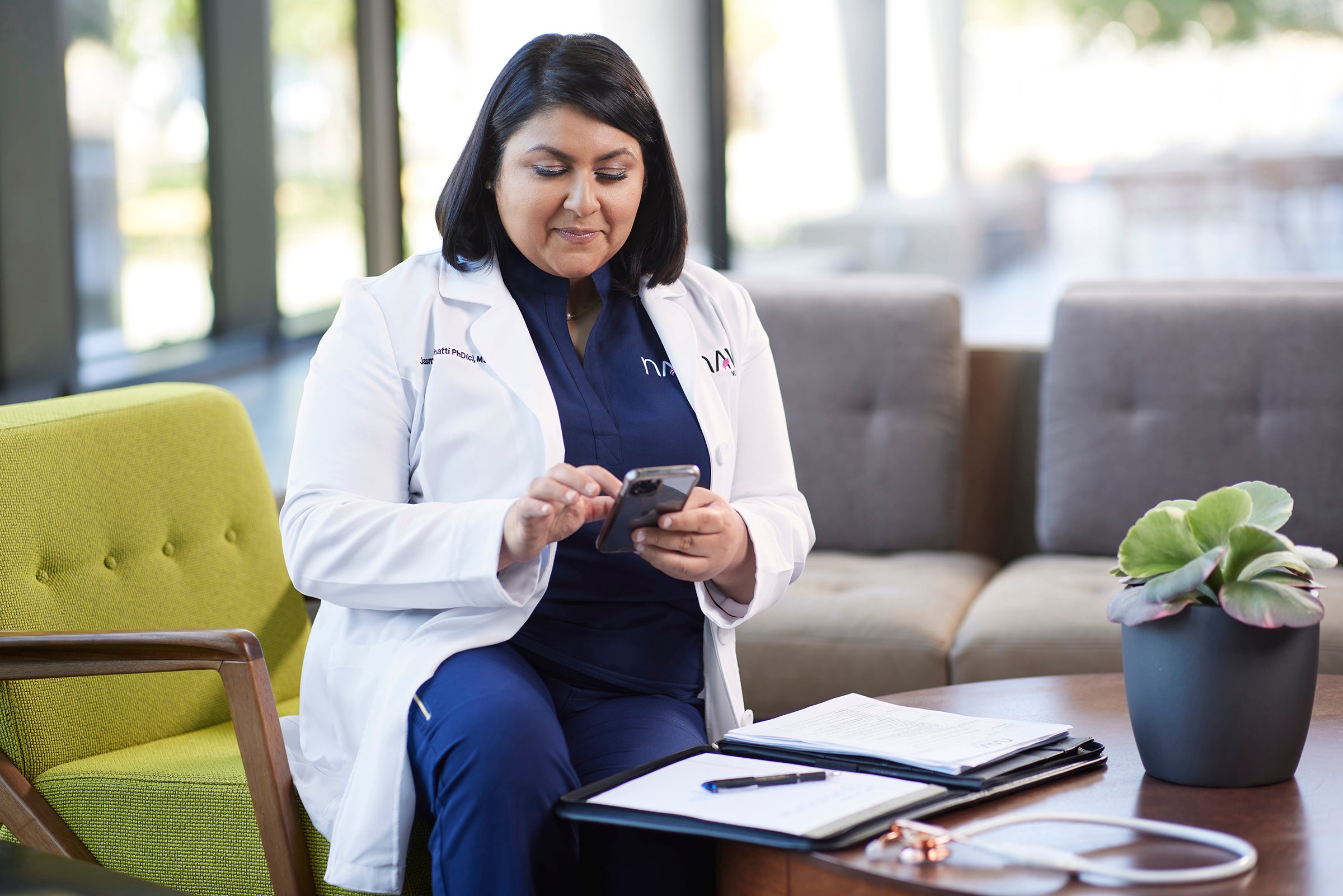 A woman in a nurse lab coat scrolls on her phone while seated at a table