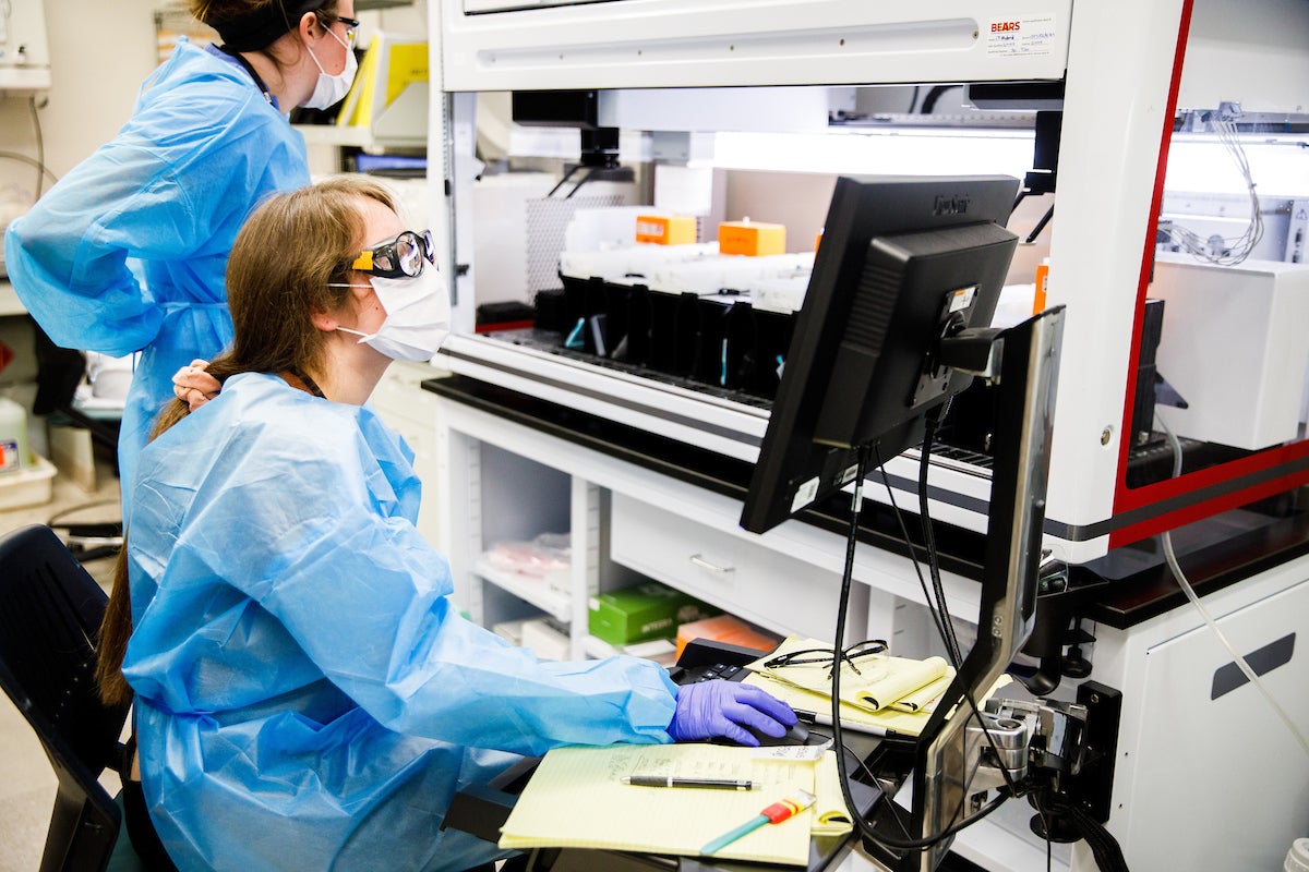 Trainees MacKenzie Wilson (left) and Gabrielle Hernandez (right) prepare an automated liquid handling robot to make KingFisher wash and elution blocks for RNA extraction.