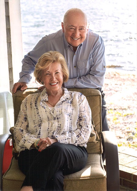 A woman sits in a chair while her husband leans on the back of it while they pose for a photo