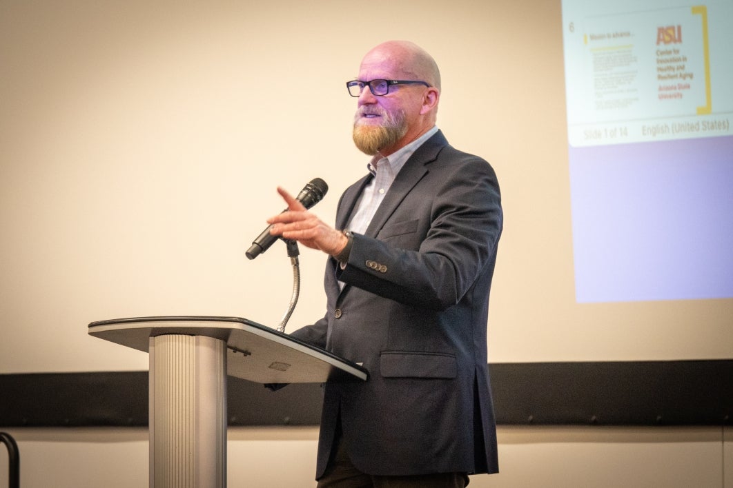 David Coon stands at a microphone in front of a powerpoint presentation. He's wearing a dark suit jacket and glasses. 