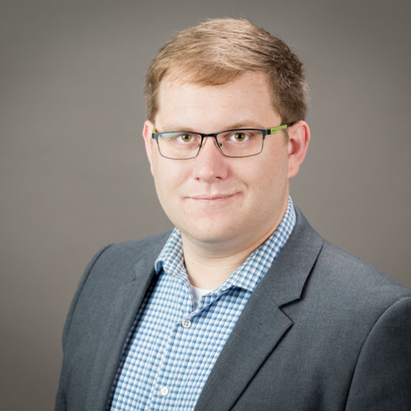  smiles with his mouth closed for a professional headshot. He's wearing a blazer with a blue and white checkered shirt underneath. His image is against a grey background