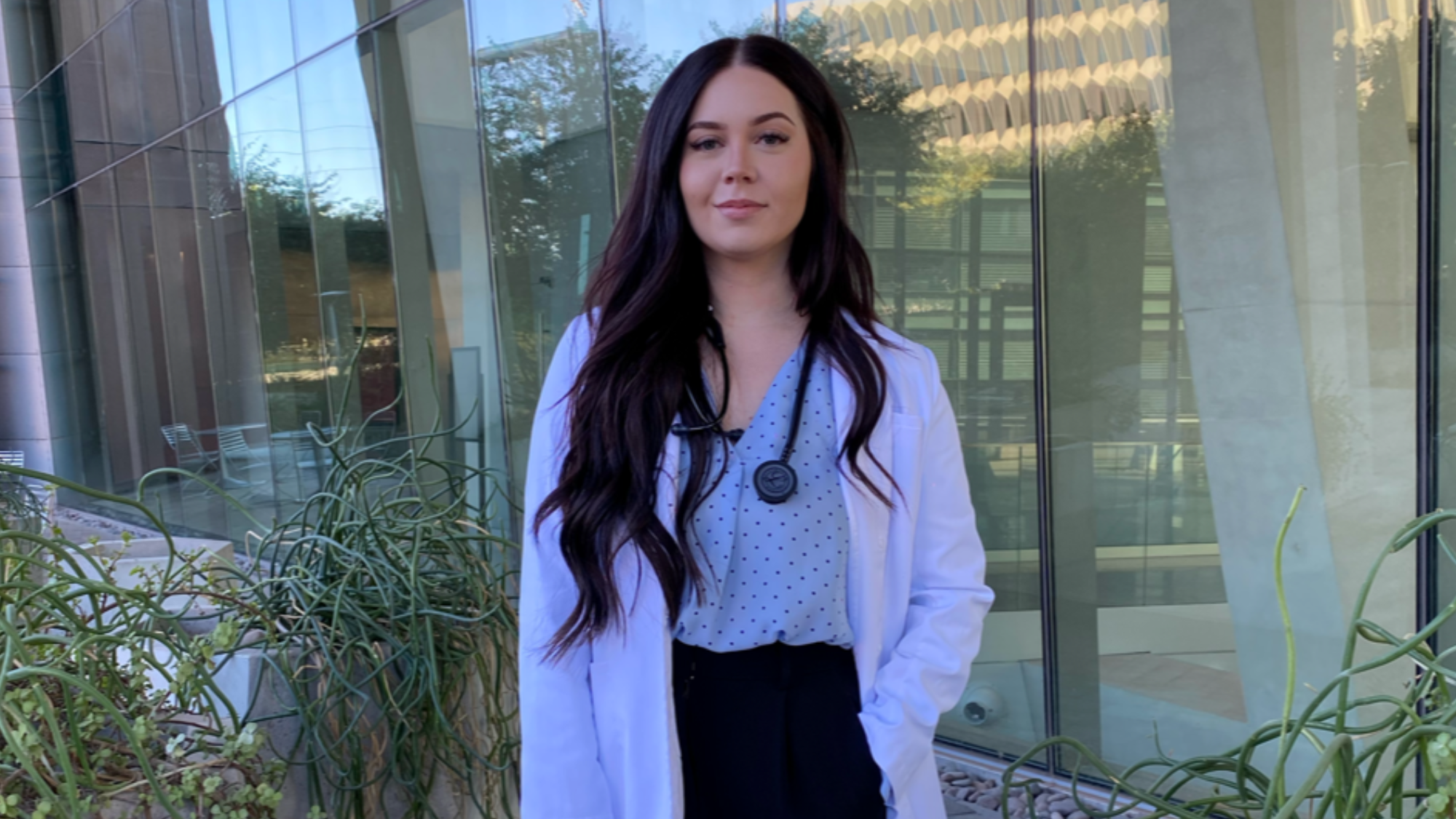  poses for the camera she is wearing a white coat and has a black stethoscope showing