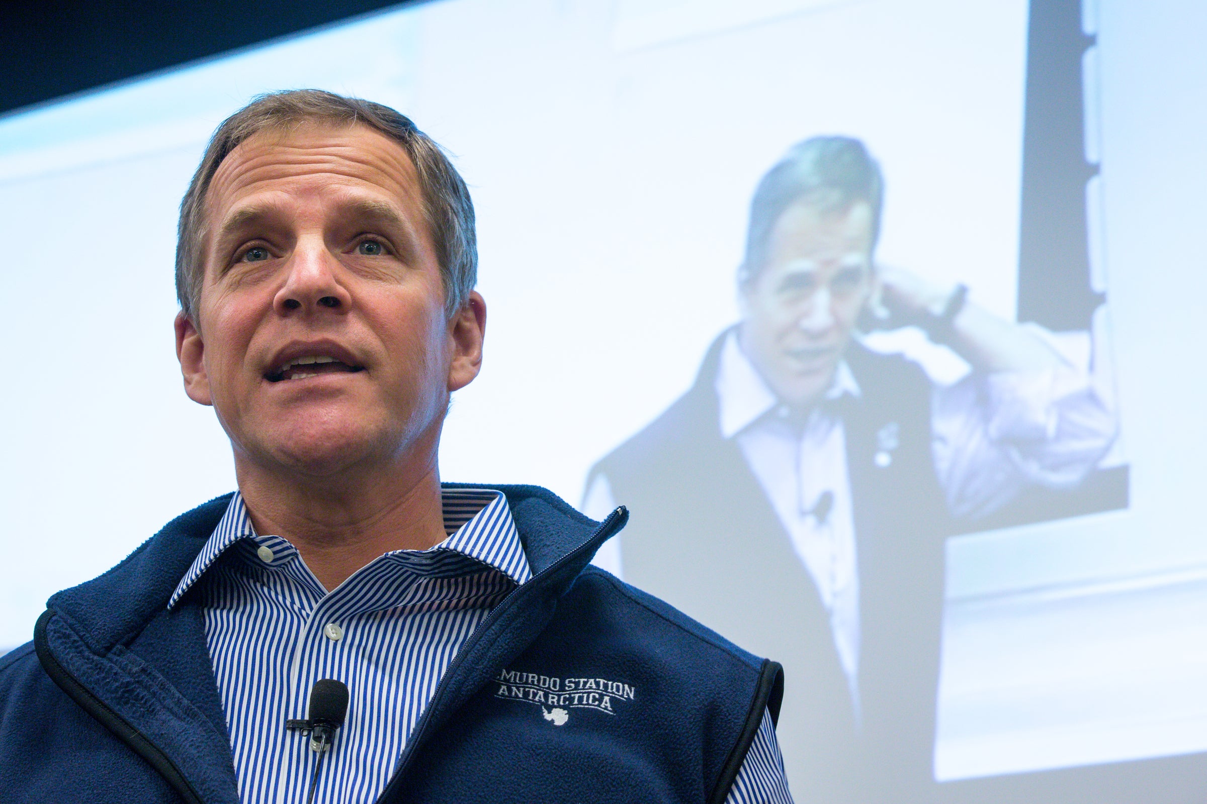 A man speaks in front of a projection screen that shows his image behind him.