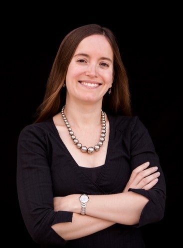  smiles at the camera. Her arms are folded across her chest and she's wearing a black blouse. She posing against a black background