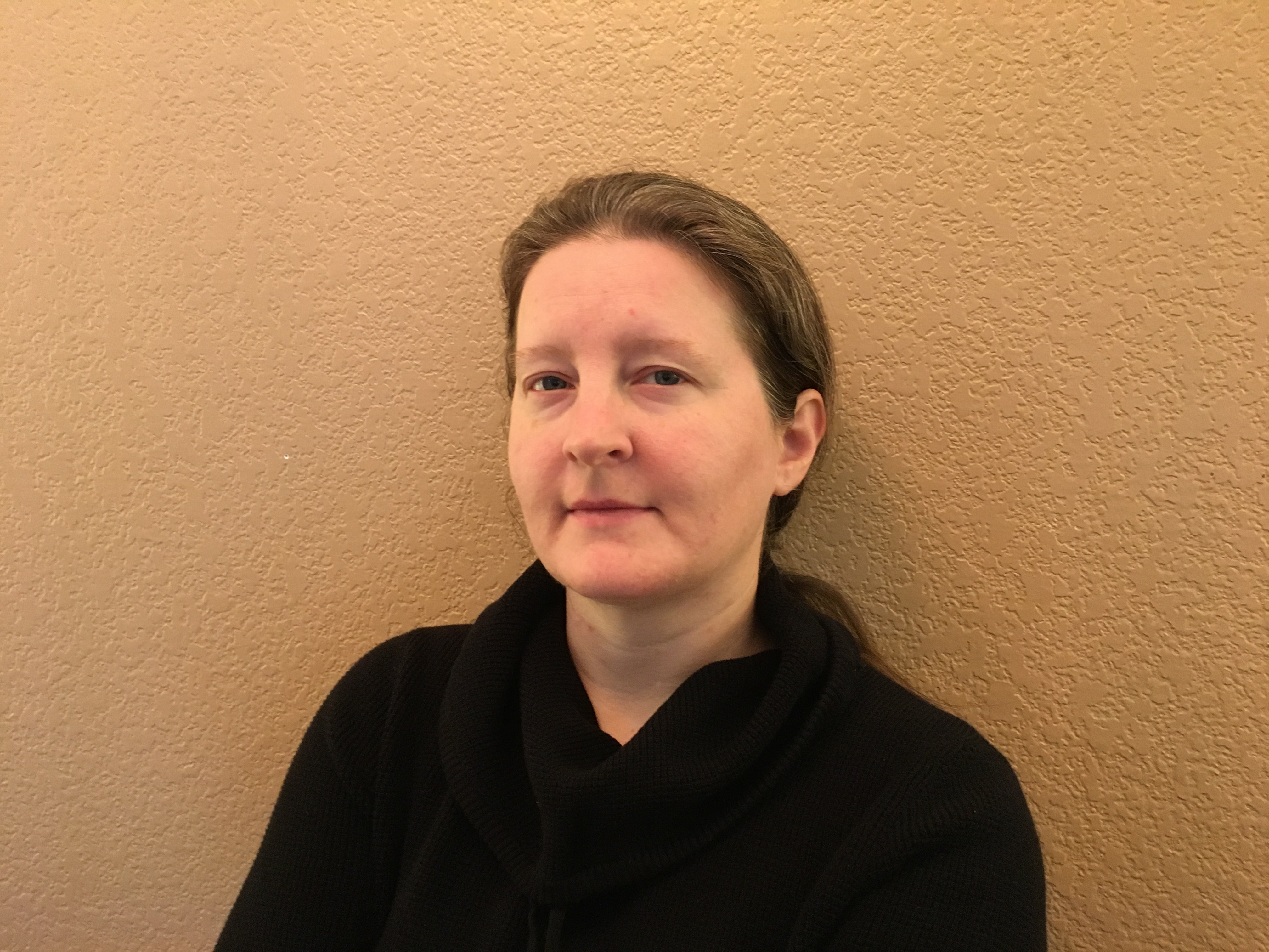 Britta Ager stands in front of a light-colored background. She is looking directly at the camera with a neutral expression. She is wearing a black top and has brown hair that is pulled back behind her ears.