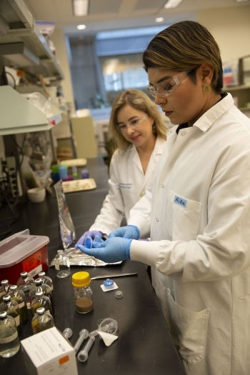 Two researchers in a lab wearing white coats, goggles and gloves as they handle specimens.