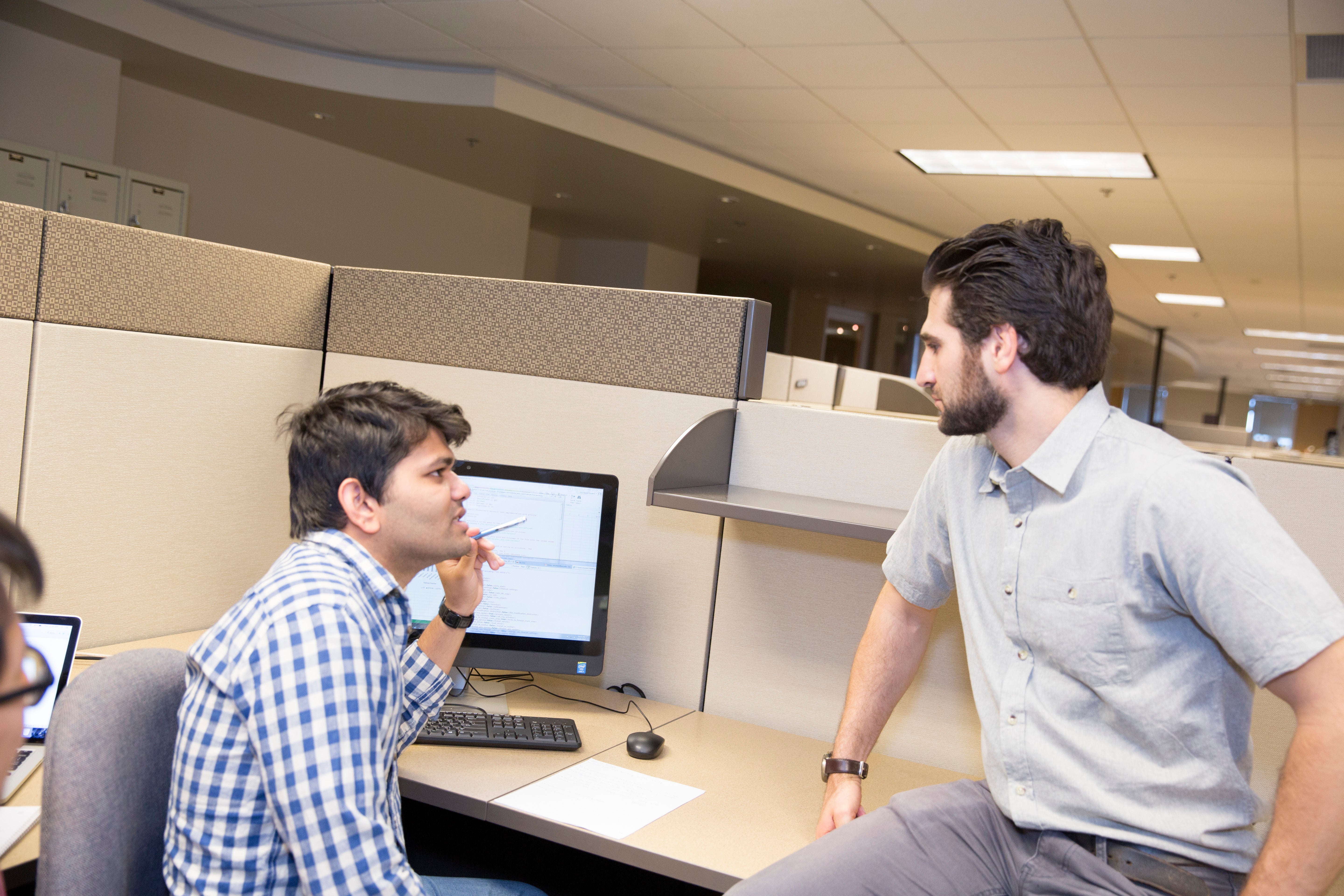 two men talking at a desk in front of a computer