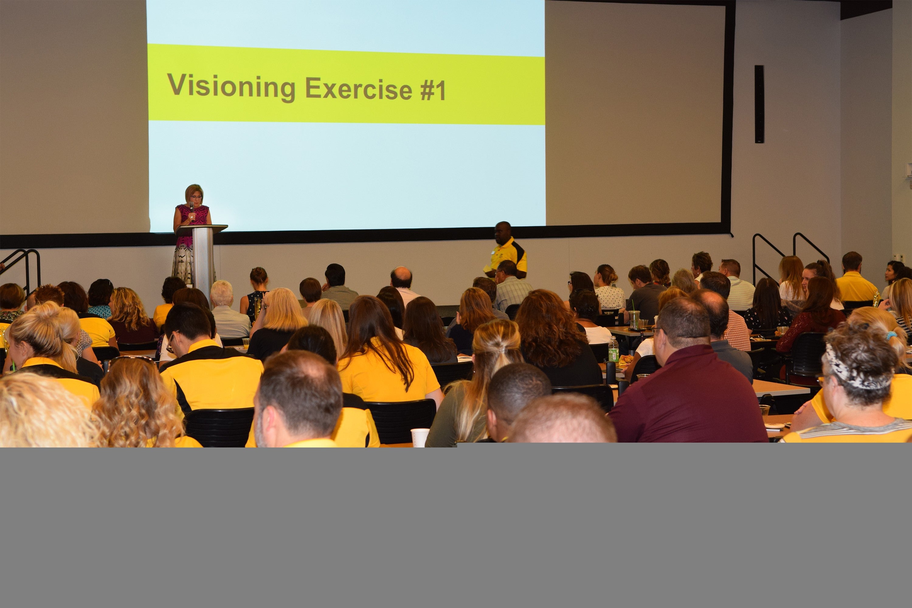 ASU College of Health Solutions Dean Deborah Helitzer stands at the front of a large auditorium, behind a lectern, next to a presentation slide that reads "Visioning Exercise #1."