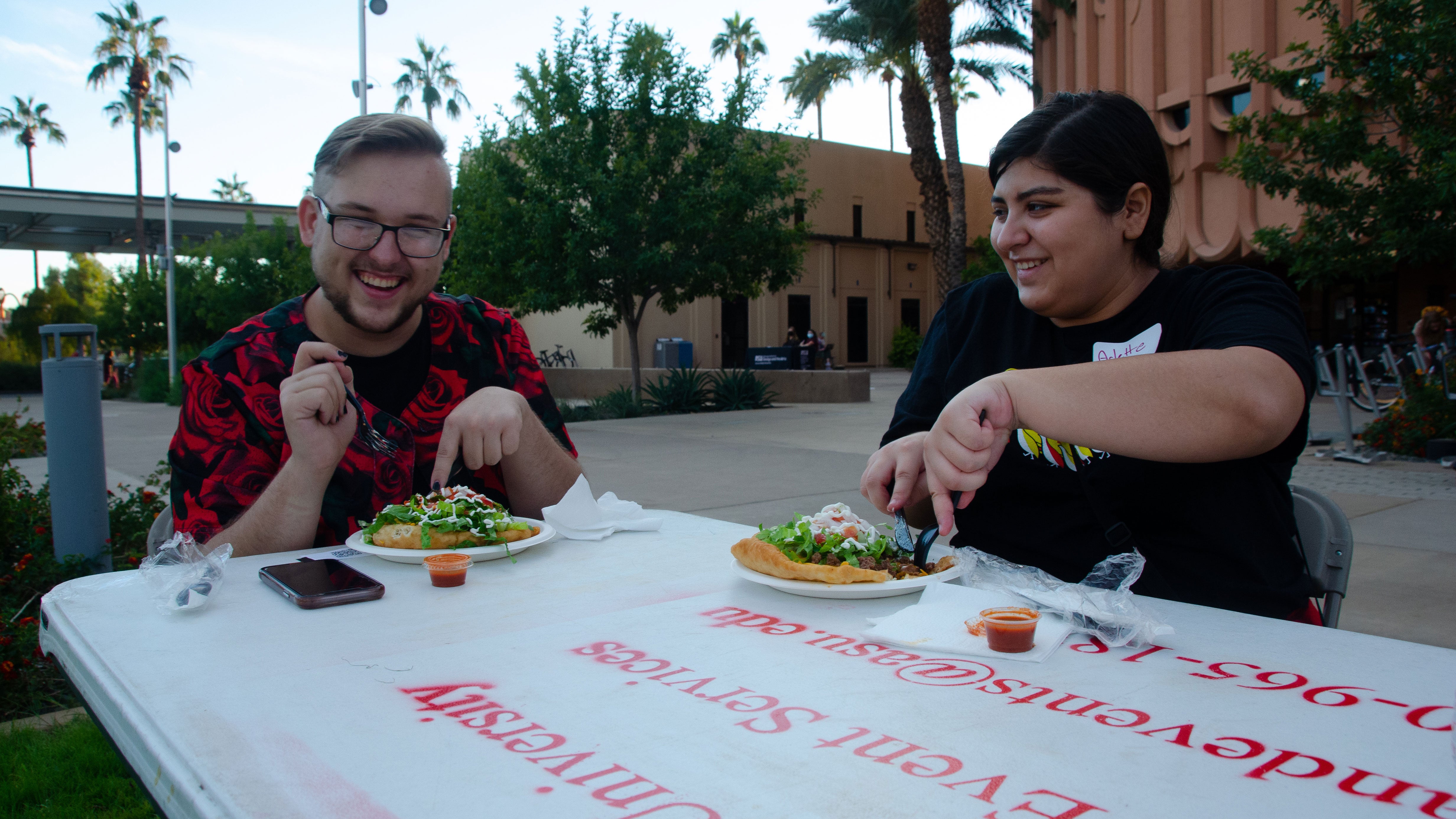 Two people sit at an outdoor table smiling while they dig into Navajo tacos