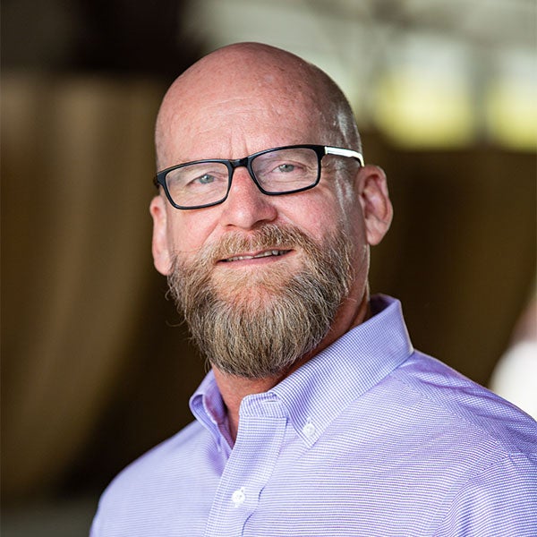  smiles at the camera for a headshot. He is wearing black rimmed glasses and a lavender shirt the background is blurred out.