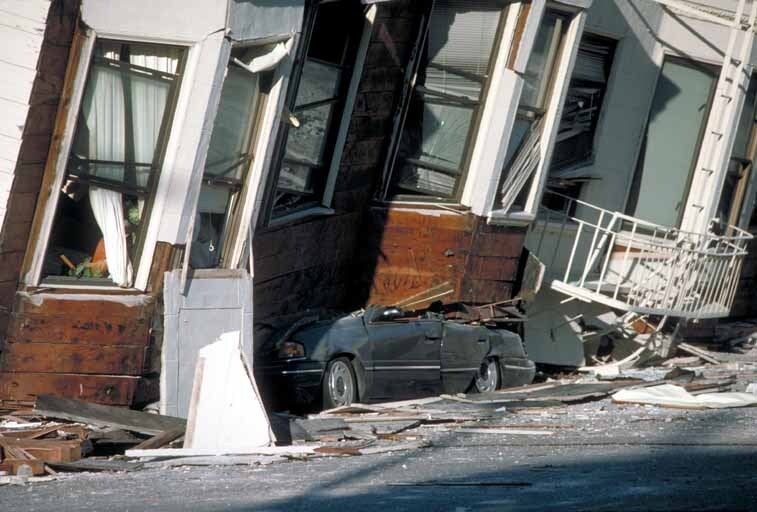 A crushed car is shown underneath a damaged building surrounded by debris.