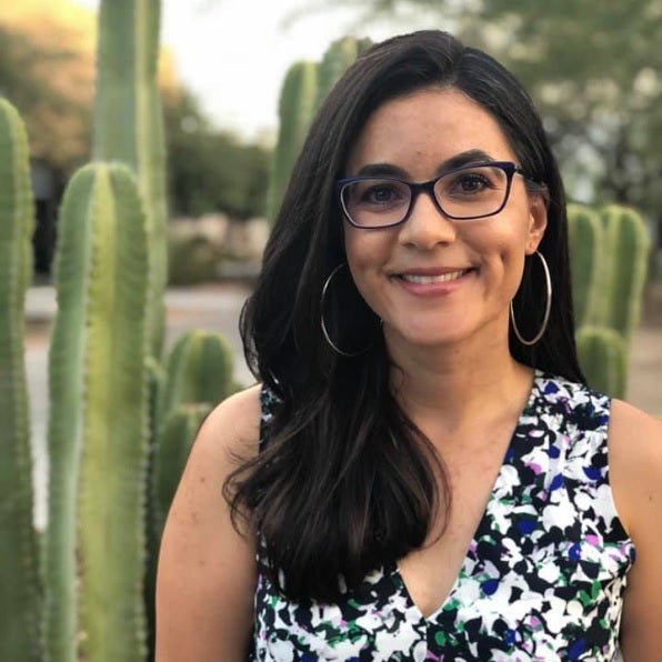 Portrait of . Fonseca-Chávez is smiling, has long, dark hair and is wearing glasses. Cacti are in the background.