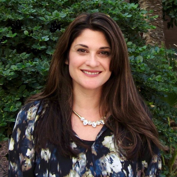 Psychology Department President's Professor  smiles straight into the camera. She's wearing a floral blouse and is standing in front of a lush green tree.