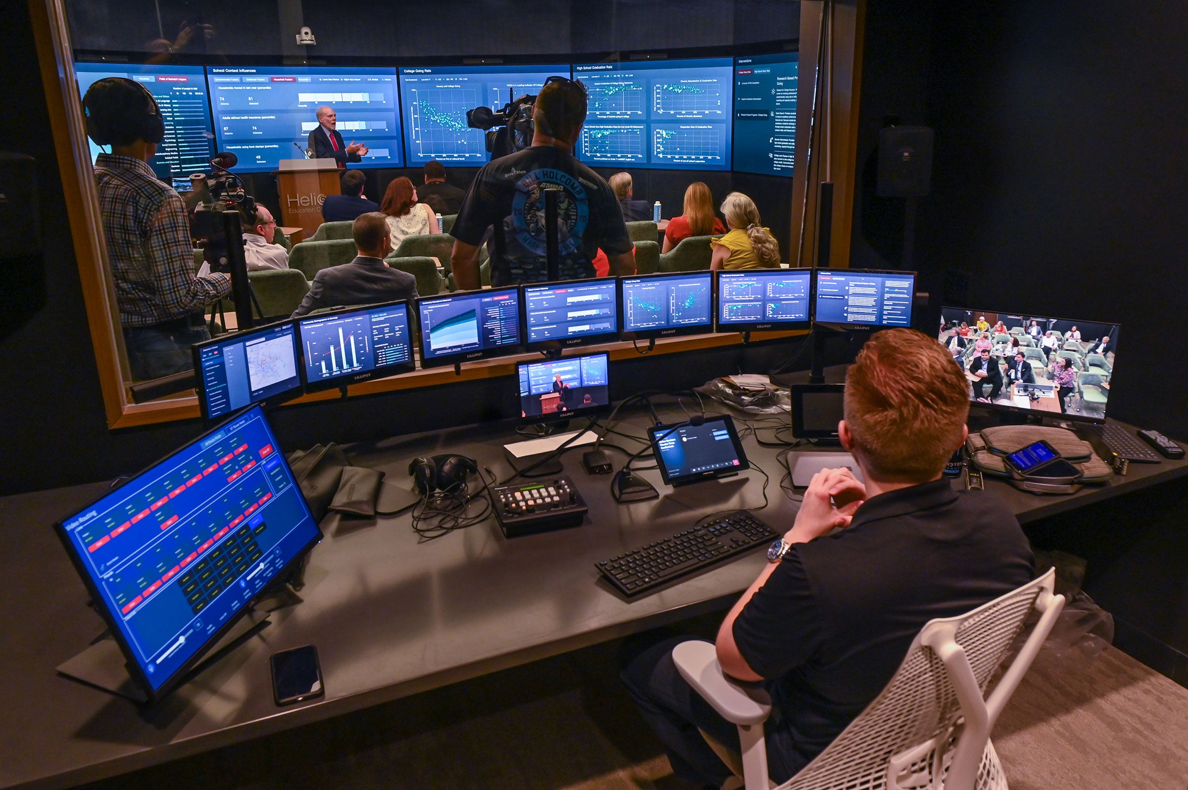 A man sits at a control desk with about a dozen screens looking into a room filled with people and a man at the front talking