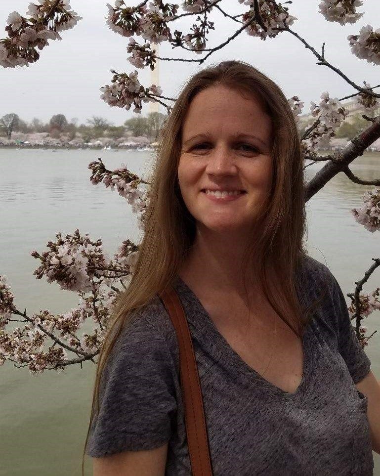  smiles at the camera. She's wearing a grey short sleeve shirt and posing in front of a tree with white flowers