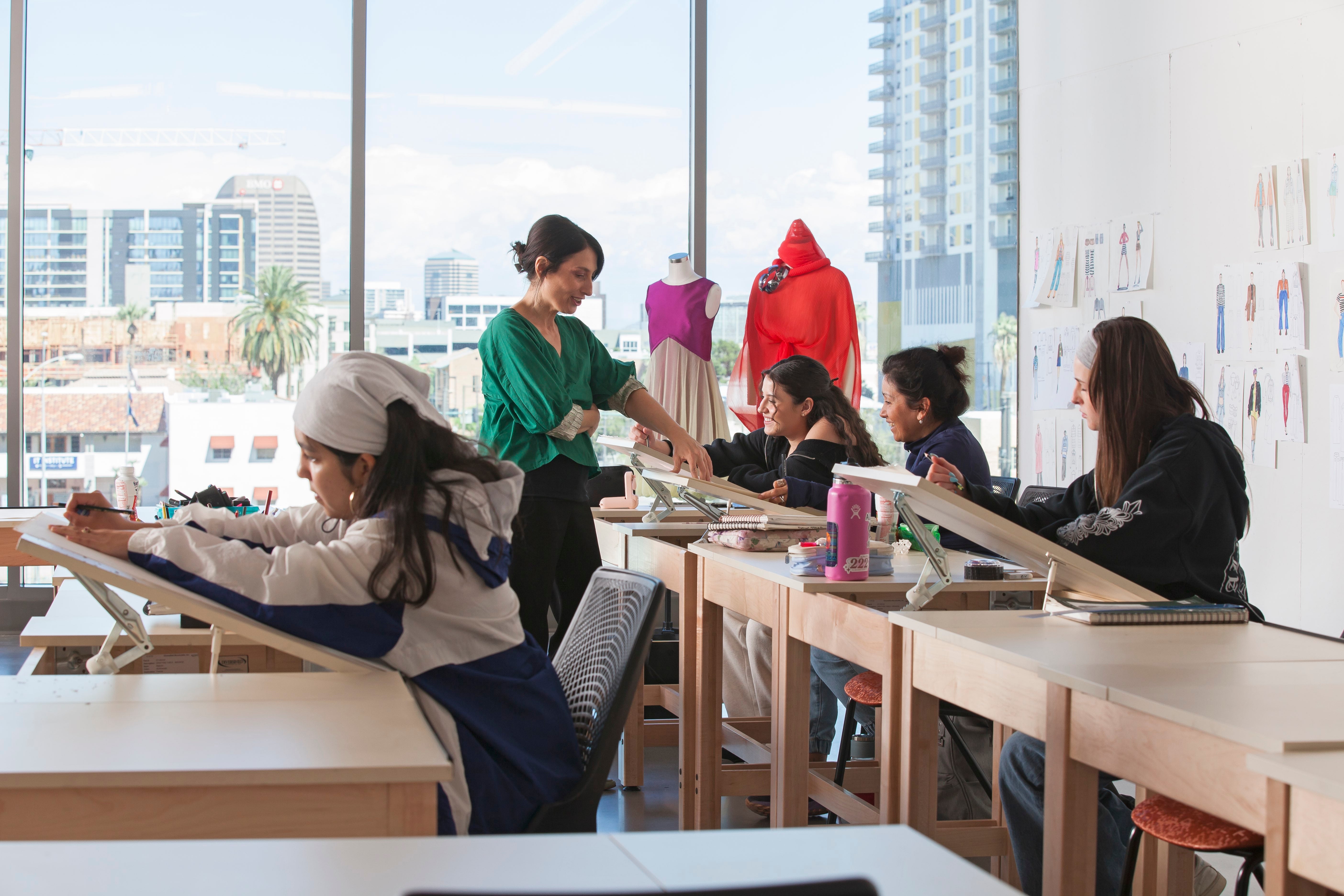 A professor works with fashion design students in a room with angled desks