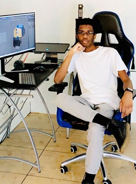 A young man sits at his computer desk, leaning his chin on his fist and looking into the camera