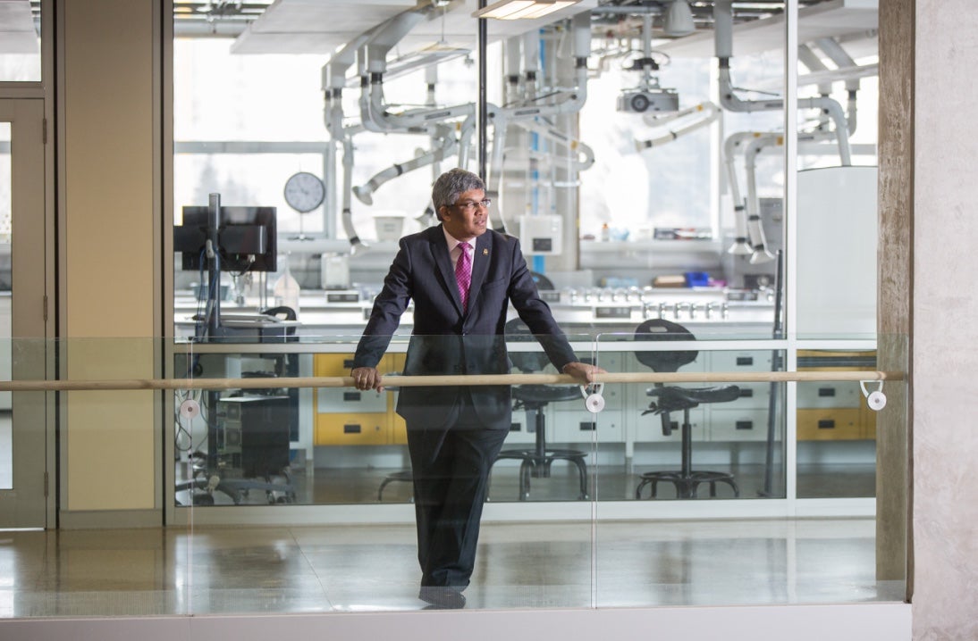 University of Calgary Professor Janaka Ruwanpura wearing a suit and leaning against a railing in front of a window overlooking a lab space.