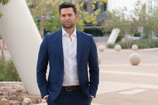  poses for a headshot outside wearing a blue suit and white dress shirt