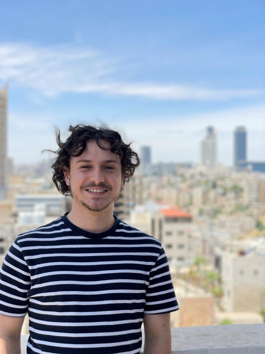 Portrait of Jordan Harb standing on a balcony with a city in the background, smiling and wearing a black and white striped shirt.