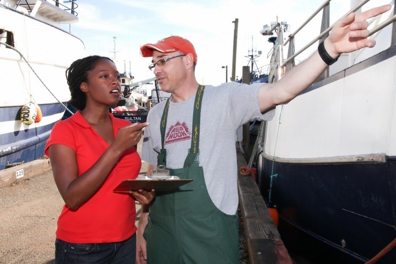 woman and man speaking on a dock with boats in the background