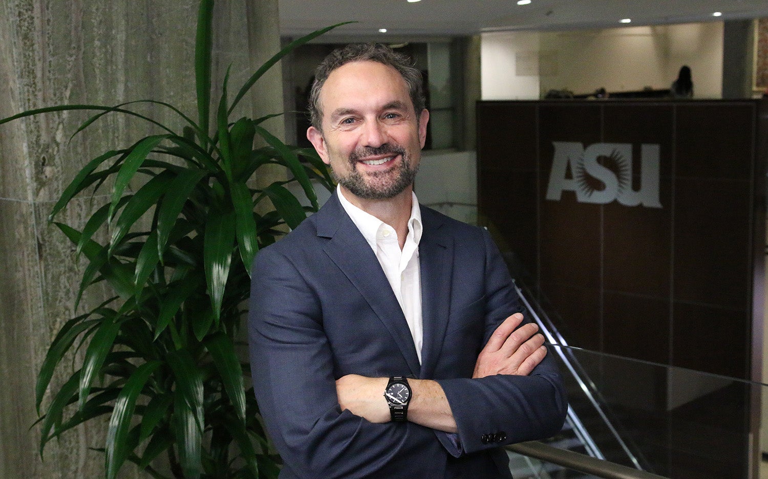  stands in gray suit, smiling with arms folded across chest on the second floor of UCENT