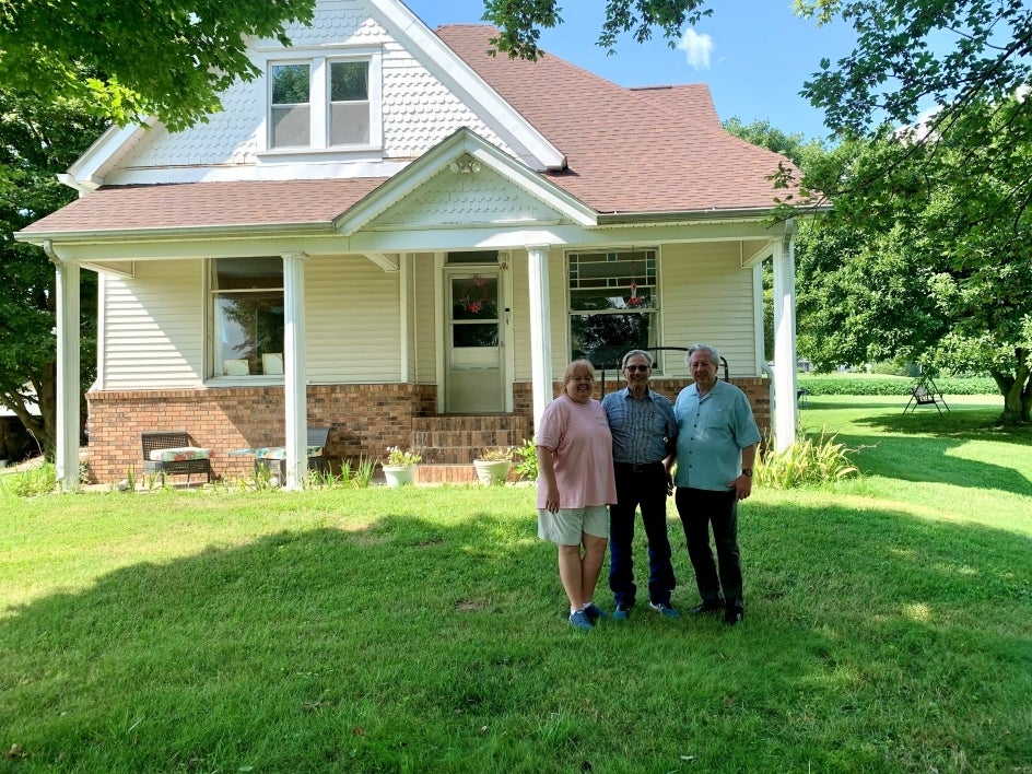 Woman and two men posing in a grassy yard in front of a white, two-story house.