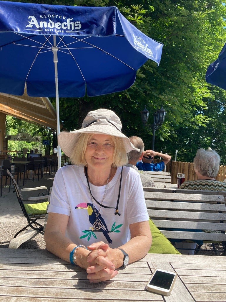 ASU anthropologist Polly Wiessner sitting outdoors at a picnic table. Wiessner smiles and wears a t-shirt with a toucan on it and a hat with a wide brim.