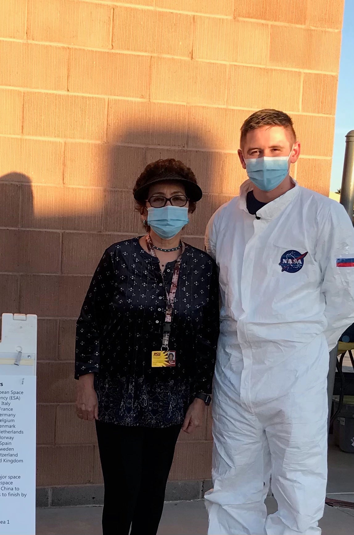 Saule Moldabekova Robb, left, and Eli Fox stand in front of a brick wall. She is wearing a dark blouse and pants and has dark hair and is wearing glasses and a blue face mask. He is wearing a mask and a white jumpsuit with a NASA logo on the breast/