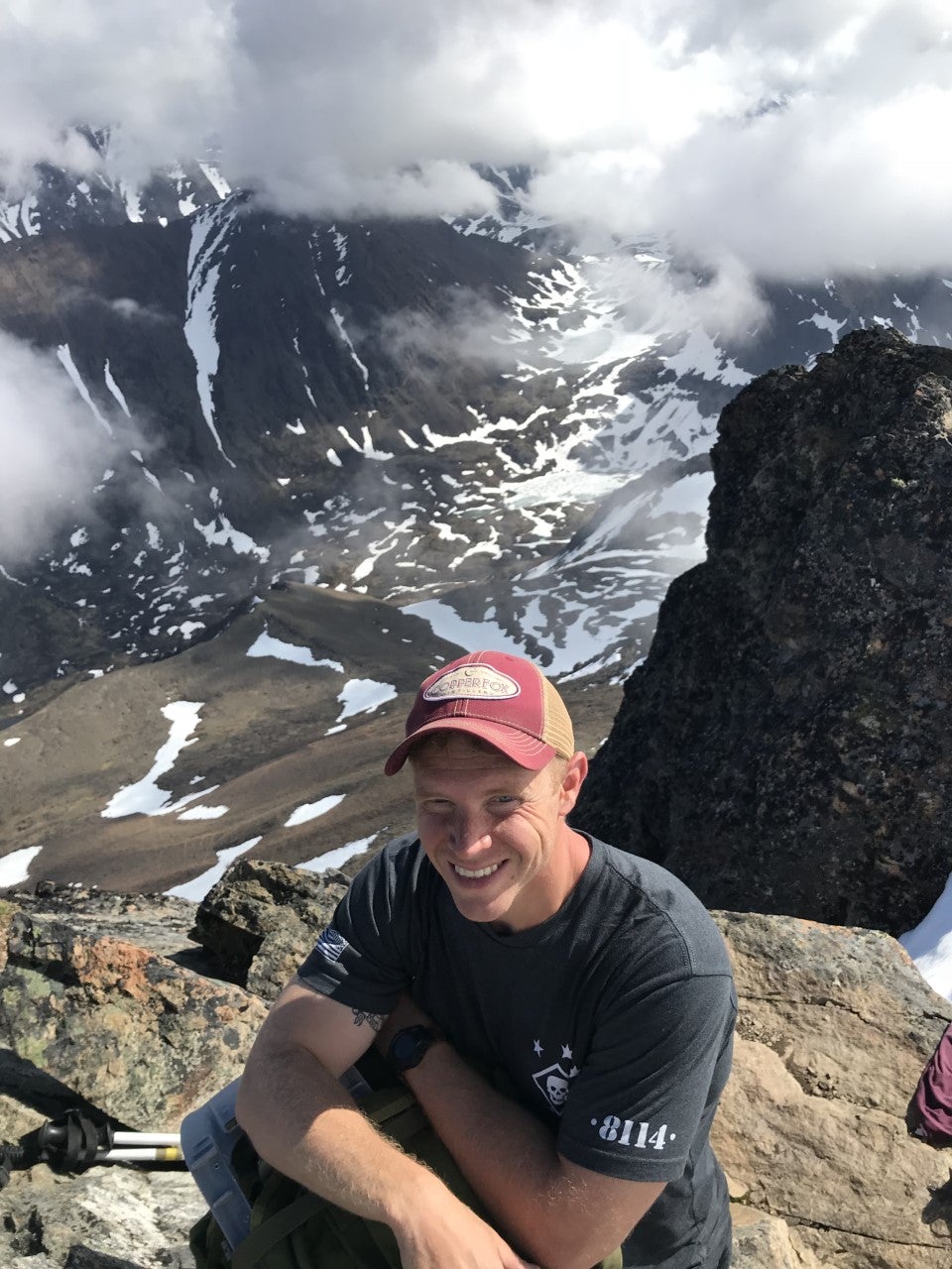 Nick sitting and smiling on a mountain peak with a snowy valley and clouds in the back